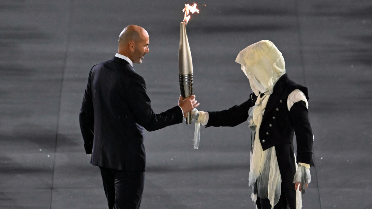 Un porteur de flambeau transmet la flamme olympique à Zinedine Zidane lors de la cérémonie d'ouverture des JO à Paris, le 26 juillet 2024.