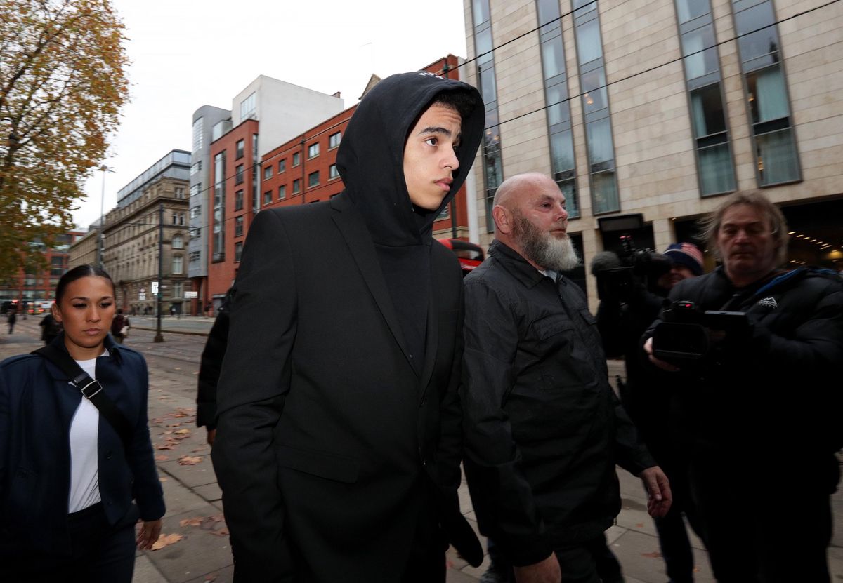 Mason Greenwood arrive au tribunal de Manchester pour son procès, vêtu d’un manteau noir à capuche, entouré de plusieurs personnes.